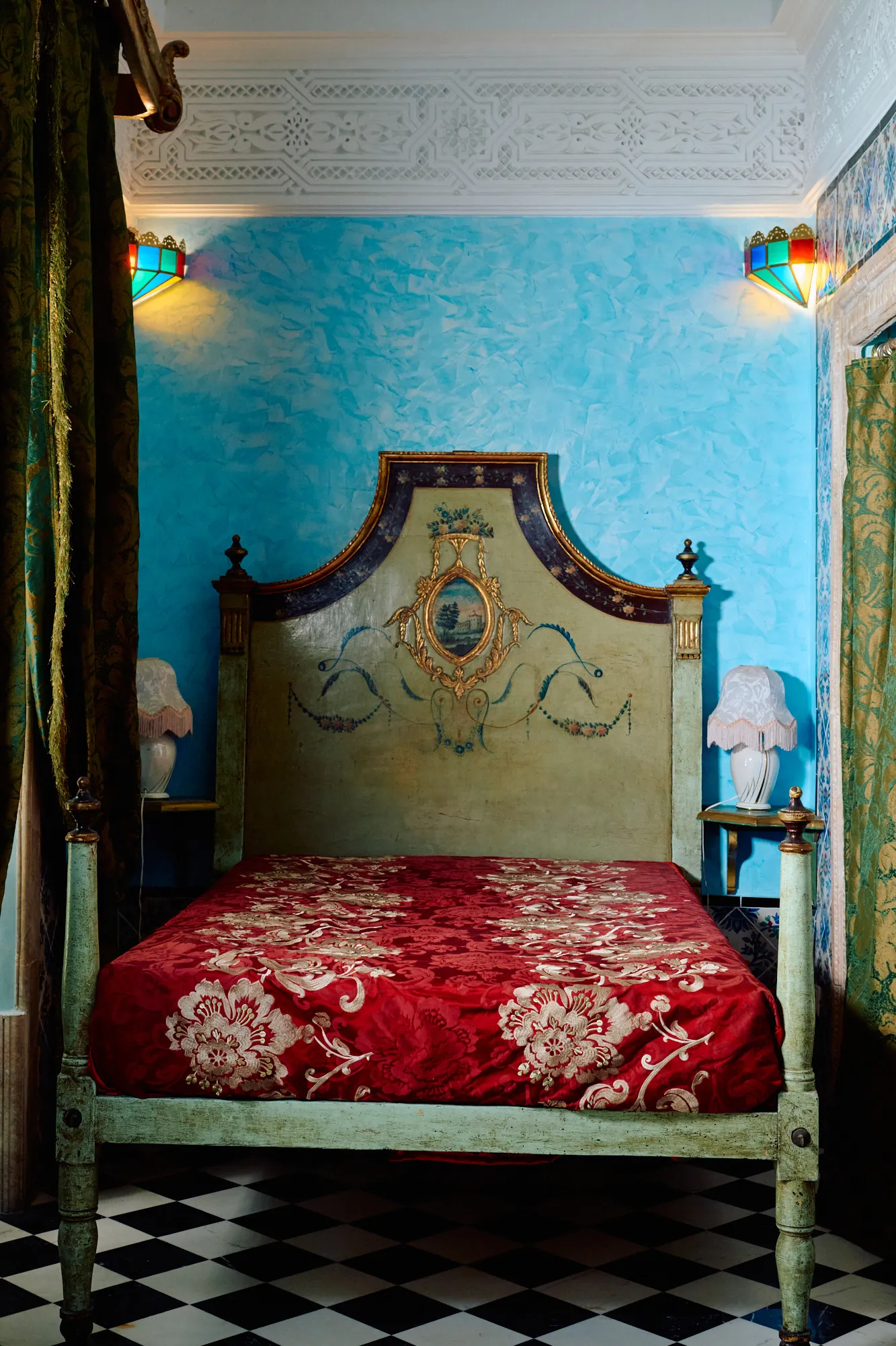 A bed with a carved headboard and a red cover, on a chequerboard floor.