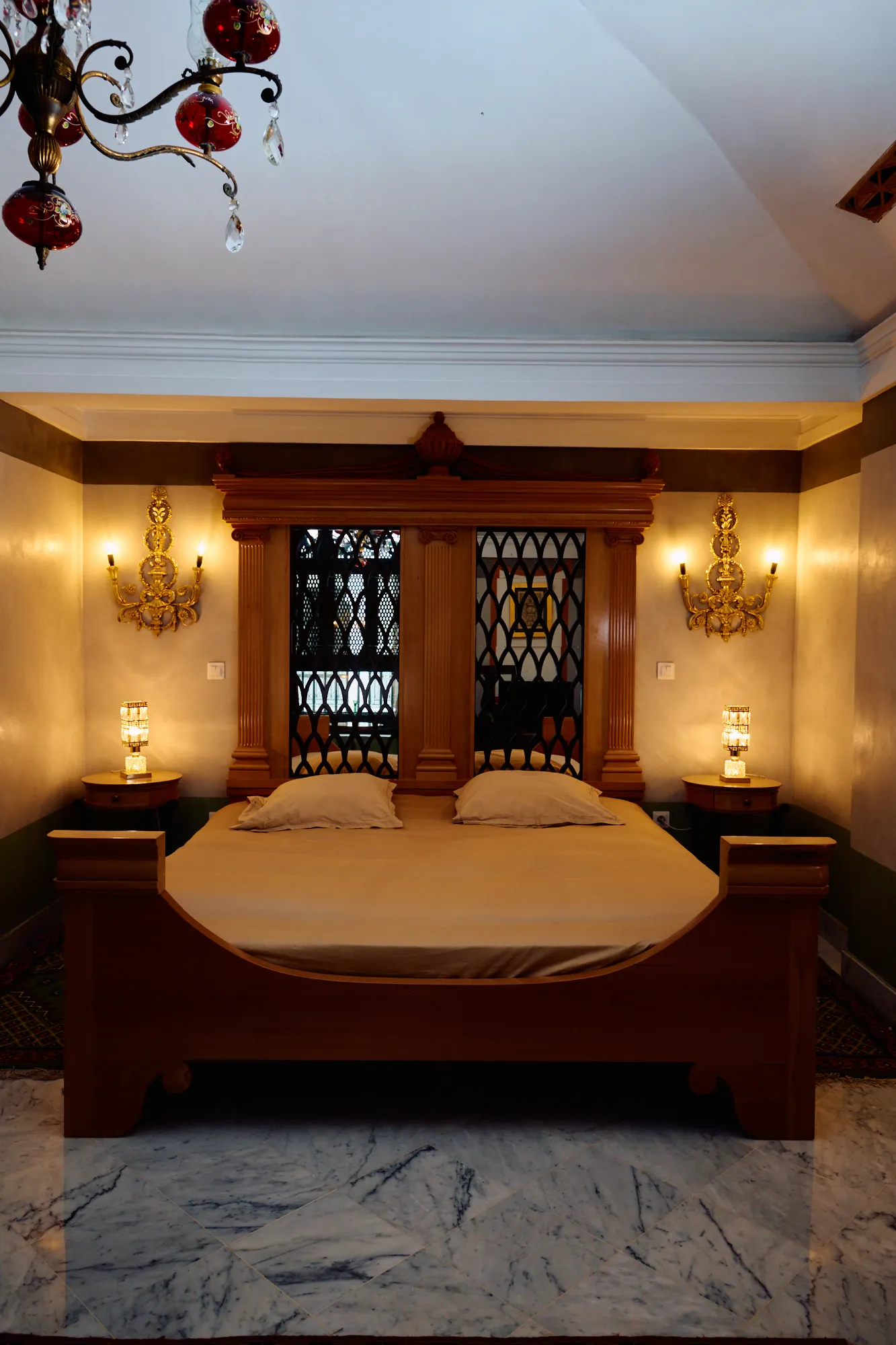 A pale bedroom with a wide bed, wall sconces and a marble floor.