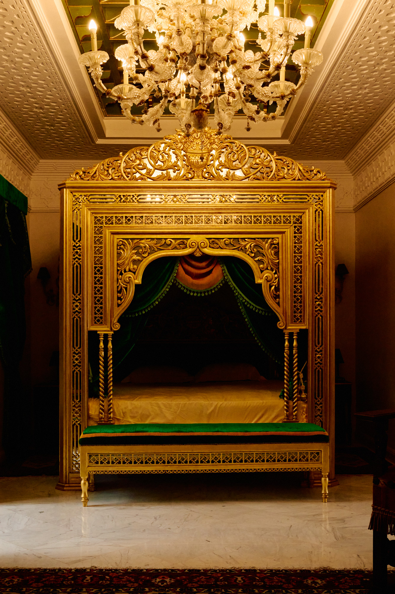 Le lit à baldaquin sous un lustre, dans un plafond de plâtre blanc sculpté.